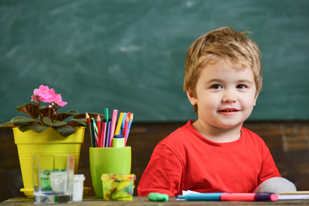 Toddler cute and cheerful ready to study. Kid boy in classroom near flower in pot and pencils, chalkboard on background, close up, copy space. Education concept. Child in school, kindergartenの写真素材