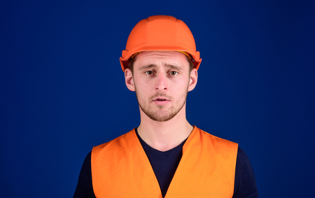 Man in protective helmet, hard hat and working uniform, blue background. Worker, contractor, builder on calm face looking at camera. Protective equipment concept. Builder in helmet posing, copy spaceの写真素材