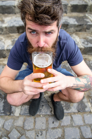Guy having rest with draught beer. Man with beard and mustache holds beer while sits on haunches, stone stairs background. Hipster on strict face drinking beer outdoor. Draught beer conceptの写真素材
