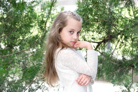 Kid model with fresh face skin in summer nature. Girl in fashion dress pose at green tree. Little child with long blond hair outdoor. Fashion beauty and hairstyle. Skincare and freshnessの写真素材