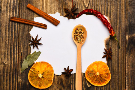 Culinary recipe concept. Piece of paper on wooden background. Spices, kitchen herbs lay around white paper. Cinnamon, dried orange and pepper, star anise lay around blank paper for recipe, copy spaceの写真素材