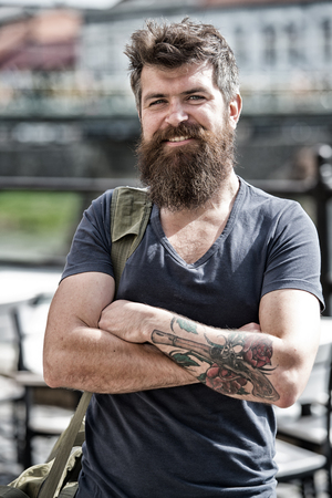 Traveller concept. Man with long stylish beard arrived at city center. Man with beard on cheerful face, urban background, defocused. Bearded man carries bag on shoulder while walkingの写真素材