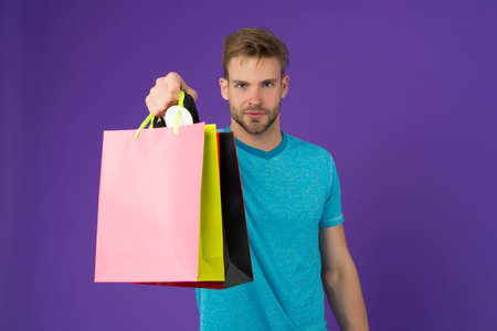 Macho with colorful paper bags on violet background. Man with shopping bags. Fashion shopper in casual blue tshirt. Holidays preparation and celebration. Shopping or sale and cyber mondayの写真素材
