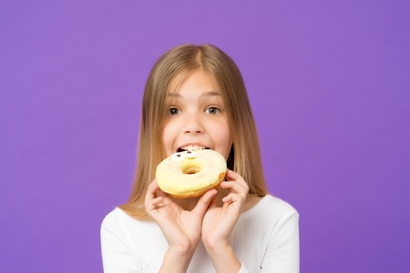 Sweetie beauty. Little girl eat donut on violet background. Child with glazed ring doughnut on purple background. Kid with junk food. Food and dessert. Childhood and childcare, copy spaceの写真素材