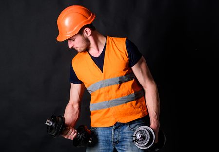 Worker, contractor, builder on busy face with muscular biceps. Builder in helmet working out, training. Man in helmet, hard hat holds heavy dumbbells, black background. Strong builder conceptの写真素材