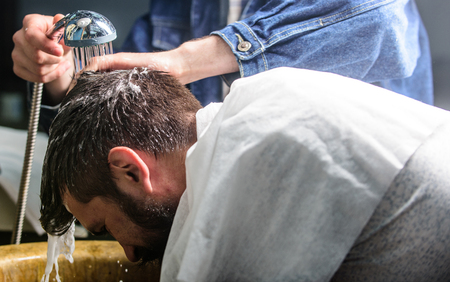 Man bearded client of hipster barbershop. Man with beard and mustache with towel on shoulders, male hands with shower on background. Barbers hands washing hair of bearded hipster. Barbershop conceptの写真素材
