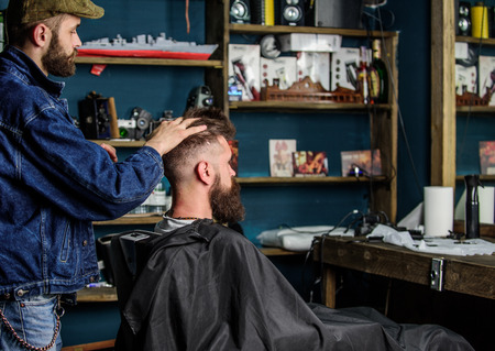 Man with beard and mustache in hairdressers chair in front of mirror, shelves on background. Barber styling hair of bearded client with wax by hands. Barbershop concept. Hipster client got hairstyleの写真素材