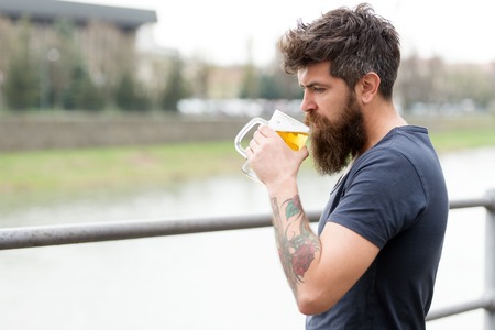 Bearded man holds beer mug, drinks beer outdoor. Man with long beard looks relaxed. Man with beard and mustache on calm face, river background, defocused. Craft beer conceptの写真素材