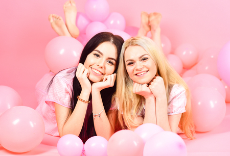 Brunette and blonde cute women in pink pajamas posing on camera while lay near air balloons at slumber party over pink background.の写真素材
