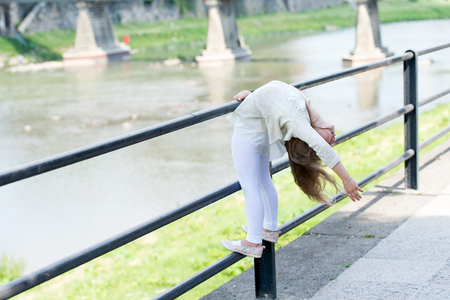 Kid girl bends backwards as flexible gymnast near railing at riverside background. Girl kid gymnast demonstrates flexibility of body. Flexibility concept. Child flexible sporty girl gymnastの写真素材