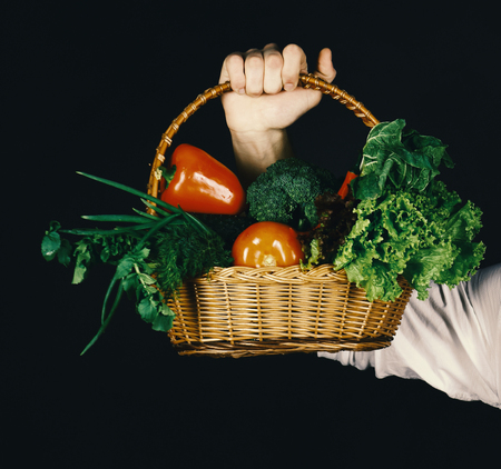 Sports and training menu. Male hand holds wicker basket with vegetables on black background.の写真素材