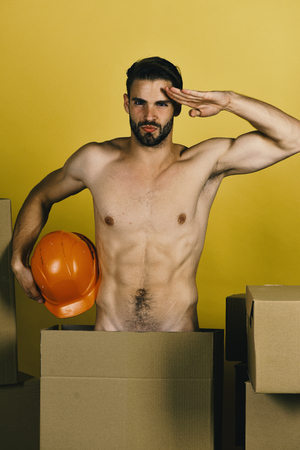 fast delivery. Guy with sexy torso saluting with hand on yellow background. Macho with bristle and strict face holding orange engineers helmetの写真素材