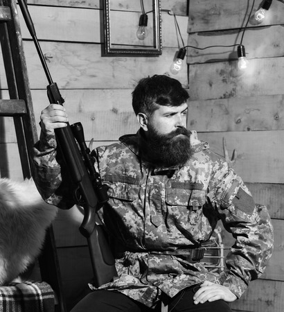 Man with beard wears camouflage clothing in wooden interior background. Hunter, brutal hipster with...
