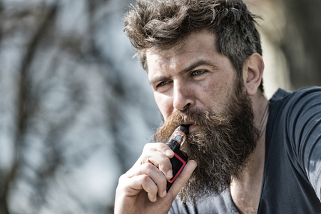 Bearded man smokes vape close up. Electronic cigarette concept. Man with long beard and clouds of smoke looks relaxed. Man with beard and mustache on calm face, branches on background, defocusedの写真素材