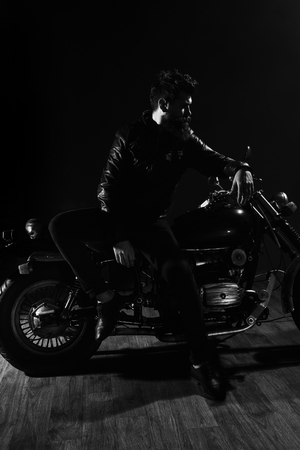 Man with beard, biker in leather jacket lean on motor bike in darkness, black background. Macho, brutal biker in leather jacket stand near motorcycle at night time, copy space. Biker culture conceptの写真素材