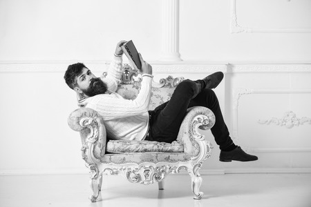 Intelligent concept. Macho smart spends leisure with book. Man with beard and mustache sits on armchair and reads book, white wall background. Scientist, professor on calm face explores literatureの写真素材