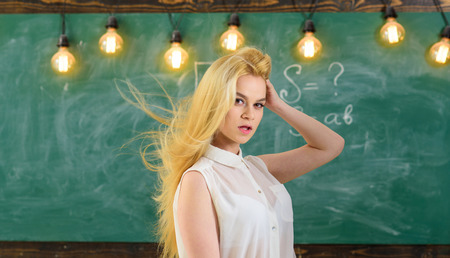 Sexy teacher concept. Woman with long hair in white blouse stands in classroom. Teacher with waving long blonde hair looks sexy. Lady strict teacher on dreamy face stands in front of chalkboardの写真素材
