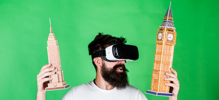 Man with beard in VR glasses, green background. Guy in VR glasses holds Big Ben and Empire State...
