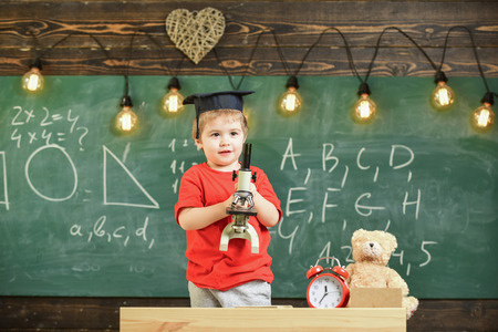 Smart kid concept. First former interested in studying, learning, education. Child on happy face holds microscope. Kid boy in academic cap work with microscope in classroom, chalkboard on backgrounの写真素材
