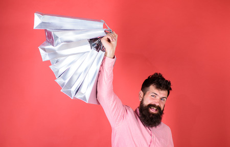 Guy shopping on sales season with discounts. Man with beard and mustache holds shopping bags, red background. Hipster on happy face is shopping addicted or shopaholic. Shopping conceptの写真素材