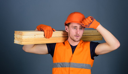 Carpenter, woodworker, strong builder on serious face carries wooden beam on shoulder. Man in helmet, hard hat and protective gloves holds wooden beam, grey background. Safety and protection conceptの写真素材