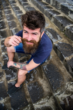 Macho with beard and mustache sitting on stone stairs background. Hipster on cheerful face relaxing outdoor. Man having rest, enjoy freshness after rain. After rain conceptの写真素材