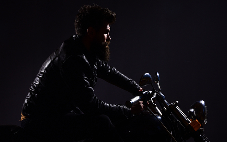 Macho, brutal biker in leather jacket riding motorcycle at night time, copy space. Man with beard, biker in leather jacket sitting on motor bike in darkness, black background. Masculinity conceptの写真素材