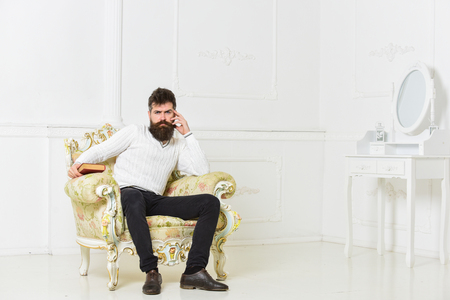 Lecturer sit on armchair and holds book, white wall background. Scientist, professor on thoughtful face analyzing literature. Man with beard and mustache spends leisure with book. Intelligent conceptの写真素材