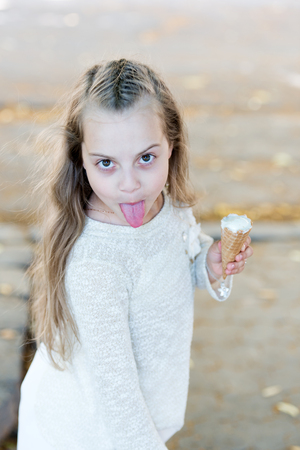 Girl sweet tooth on cheerful grimace with tongue eats ice cream, light background. Kid girl with ice cream cone in hand. Summer treats concept. Sweet tooth child with white ice cream in waffle coneの写真素材
