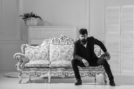 Fashion and style concept. Macho attractive and elegant on serious face and thoughtful expression. Man with beard and mustache wearing fashionable classic suit, sits on old fashioned couch or sofaの写真素材