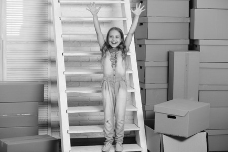 Kid stands on white ladder and puts her hands up. Child near cardboard boxes in pile. Childhood and new homeの写真素材