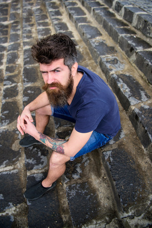 Man with beard and mustache sitting on stone stairs background. Hipster on strict face relaxing outdoor. After rain concept. Guy having rest, enjoy freshness after rainの写真素材
