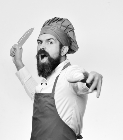 Cook with angry face in burgundy apron and chef hat. Chef pretends to throw blue knife. Cooking and professional culinaryの写真素材