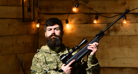 Man with beard wears camouflage clothing in wooden interior background. Macho on strict face at...
