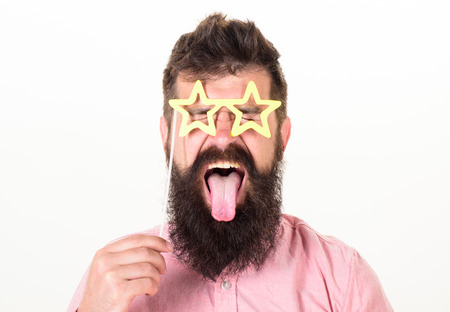 Man with stickout tongue, closed eyes and beard wearing paper star shaped glasses, fun concept. Hipster with crazy look and long beard on white background. Bearded man posing with party accessoriesの写真素材