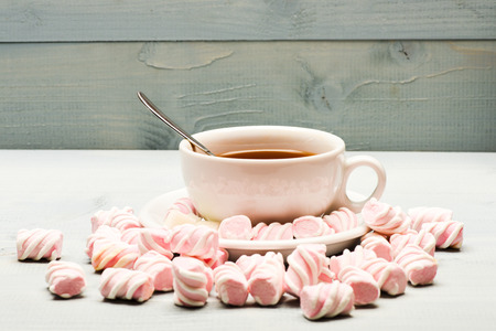 Cup with hot black tea and marshmallows. Cup and sweets prepared as dessert. Tea break concept. Mug filled with black brewed tea, spoon and heap of marshmallow on white wooden backgroundの写真素材