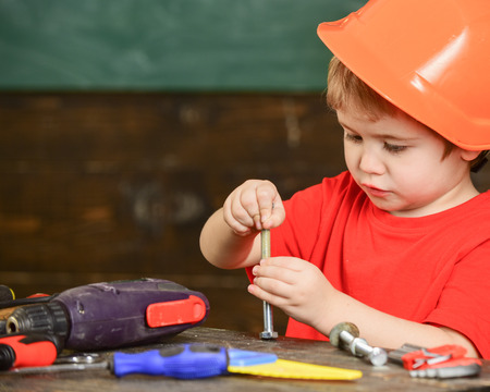 Toddler on busy face plays with bolts at home in workshop. Handcrafting concept. Child in hard hat, helmet playing with hex bolts as builder or repairer, handcrafting. Kid boy play as handymanの写真素材