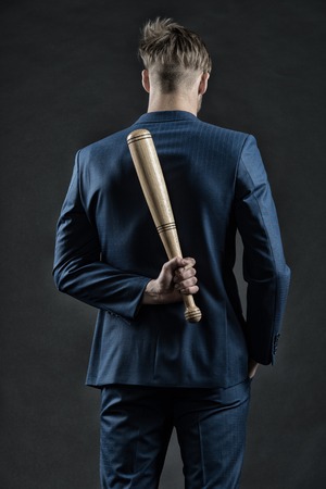Latent aggression. Man with bat hides his aggression slow down and keep calm, rear view. Businessman or man in formal suit hides wooden bat behind back, dark background. Hidden danger conceptの写真素材