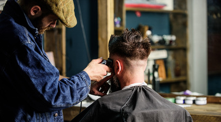 Barber with clipper trimming hair of client, rear view. Working process concept. Barber with hair clipper works on short haircut on teple, barbershop background. Hipster client getting short haircutの写真素材