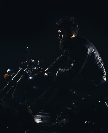 Bikers leisure concept. Macho, brutal biker in leather jacket riding motorcycle at night time, copy space. Man with beard, biker in leather jacket sitting on motor bike in darkness, black backgroundの写真素材