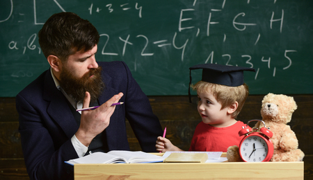 Happy family. Father and son doing homework together. Teacher in formal wear and pupil in mortarboard in classroom, chalkboard on background. Father teaches son, discuss, explain. Education conceptの写真素材