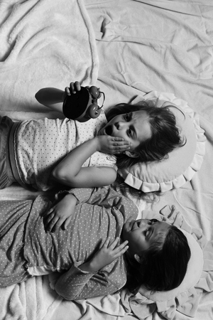 Schoolgirls in pink pajamas lie on colorful pillows, top view. Girls lie on white and pink sheets holding red clockの写真素材