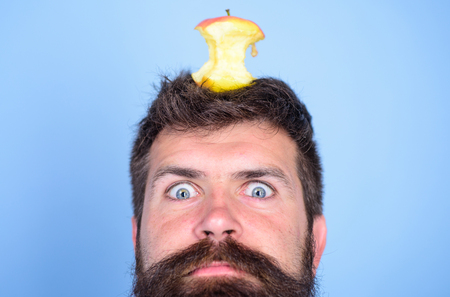 Hipster shocked face with apple stump target on head blue background, close up. Man handsome hipster long beard almost eaten apple stump on head as target. Weight loss goal. Live target conceptの写真素材