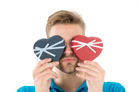 Guess which one. Man holds two heart shaped gift boxes in front of eyes, white background. Macho prepared romantic surprise for valentines day. Valentines day gift. Romantic surprise for herの写真素材