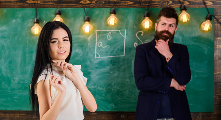 Man with beard and young lady teacher stand in classroom, chalkboard on background. Lady teacher and...