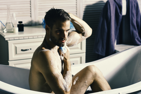 Sexuality and healthcare concept. Guy in bathroom with toiletries on background. Man with beard and confident look taking bath. Macho sitting naked in bathtub washing with sponge and showing muscles.の写真素材