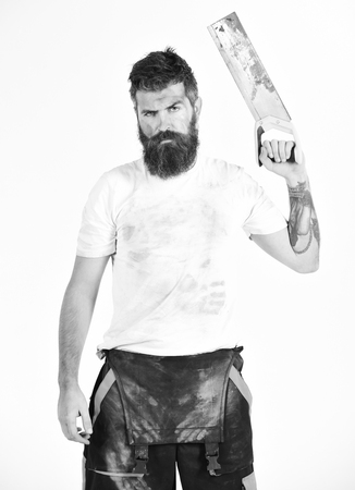 Builder, worker, carpenter, handyman raised hand with saw, white background. Man on strict face in dirty shirt holds handsaw. Guy with beard and mustache covered with dust. Dusty work conceptの写真素材