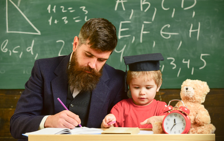 Busy kid studying at school. Teacher, father checking homework, helps to boy, son. Teacher in formal wear and pupil in mortarboard in classroom, chalkboard on background. Individual studying concept.の写真素材