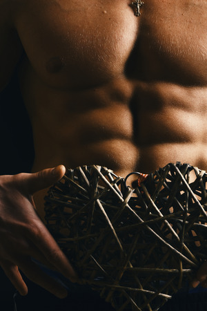 Sports, bodybuilding, love and decor concept. Guys abs and strong chest. Decoration and male with naked torso on background, defocused. Athletes body in close up, hand holding big rattan heart.の写真素材