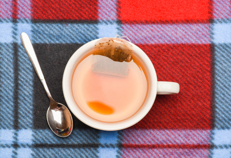 Tea brew concept. Mug filled with boiling water, teabag and spoon on colorful plaid background. Process of tea brewing in ceramic mug, top view. Mug filled with hot water and dipped bag of tea.の写真素材